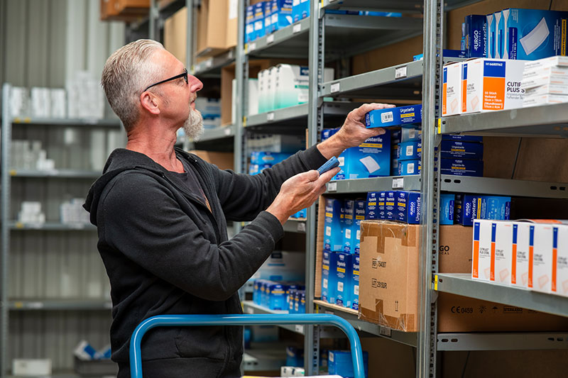 Dieses Bild zeigt einen Mitarbeiter in einem medizinischen Lager, der medizinische Produkte wie Wundverbände der Marken Urgo und Smith & Nephew (Cutiplast) kommissioniert.